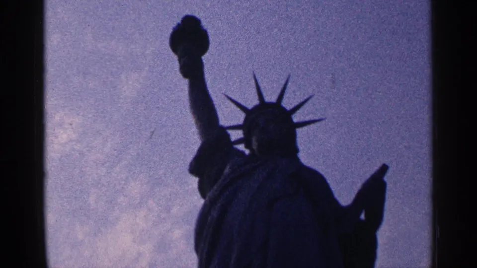 1964 New York City 8mm frame showing the Statue of Liberty in dark silhouette against a purple-blue sky.