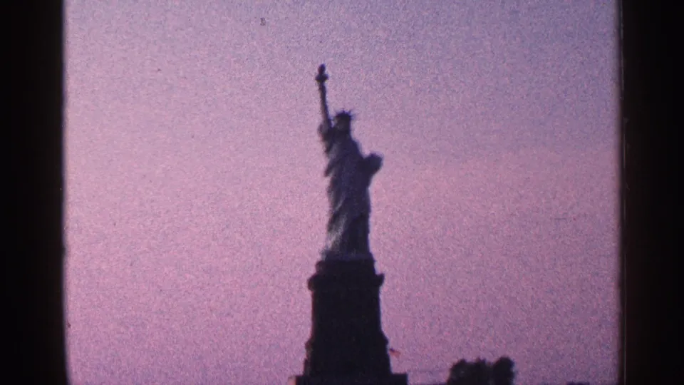 1964 8mm New York City home-movie frame showing the full Statue of Liberty and pedestal as a dark silhouette against a purple twilight sky.