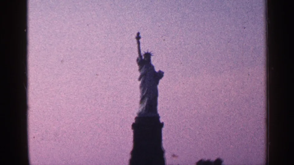 1964 8mm New York City home-movie frame with the Statue of Liberty and pedestal silhouetted in the distance under a muted purple sky.