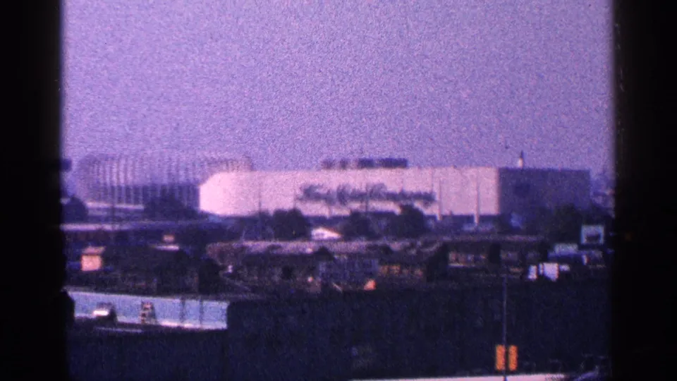 Soft 1964 New York City frame with a large light-colored venue-like building seen in the distance through grain and haze.