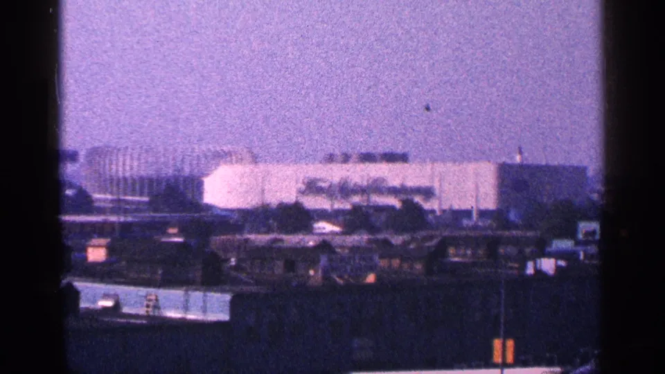 Purple-tinted 8mm frame of a distant New York City building complex, with low foreground structures and heavy film grain.