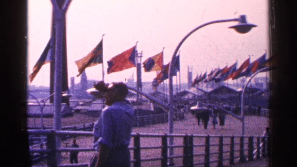 1964 New York City home-movie frame showing visitors near a railing, with rows of international flags and curved streetlamps in view.