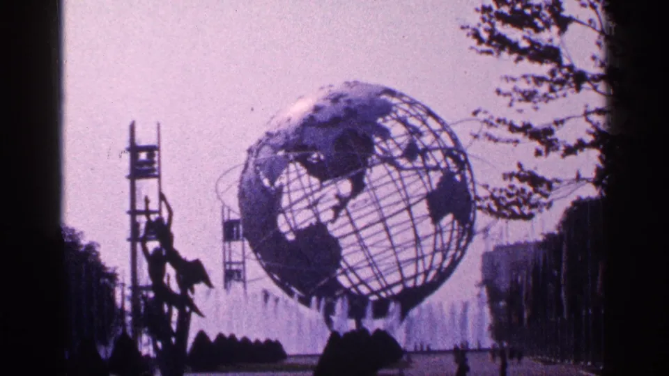 1964 8mm view of the Unisphere at Flushing Meadows Corona Park, framed across fountain spray under a hazy sky.