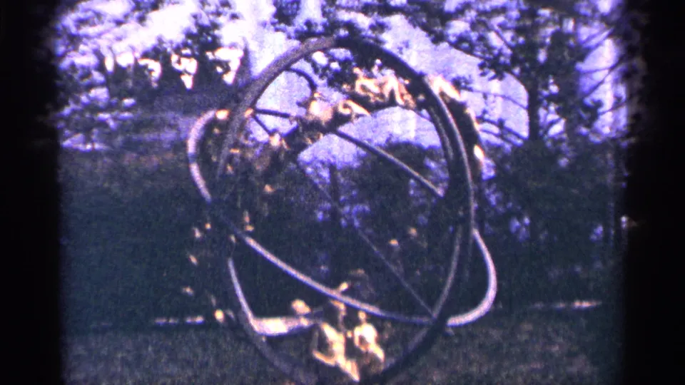 Vintage 8mm frame from Flushing Meadows Corona Park showing a ringed metal sculptural form, trees, and grass.