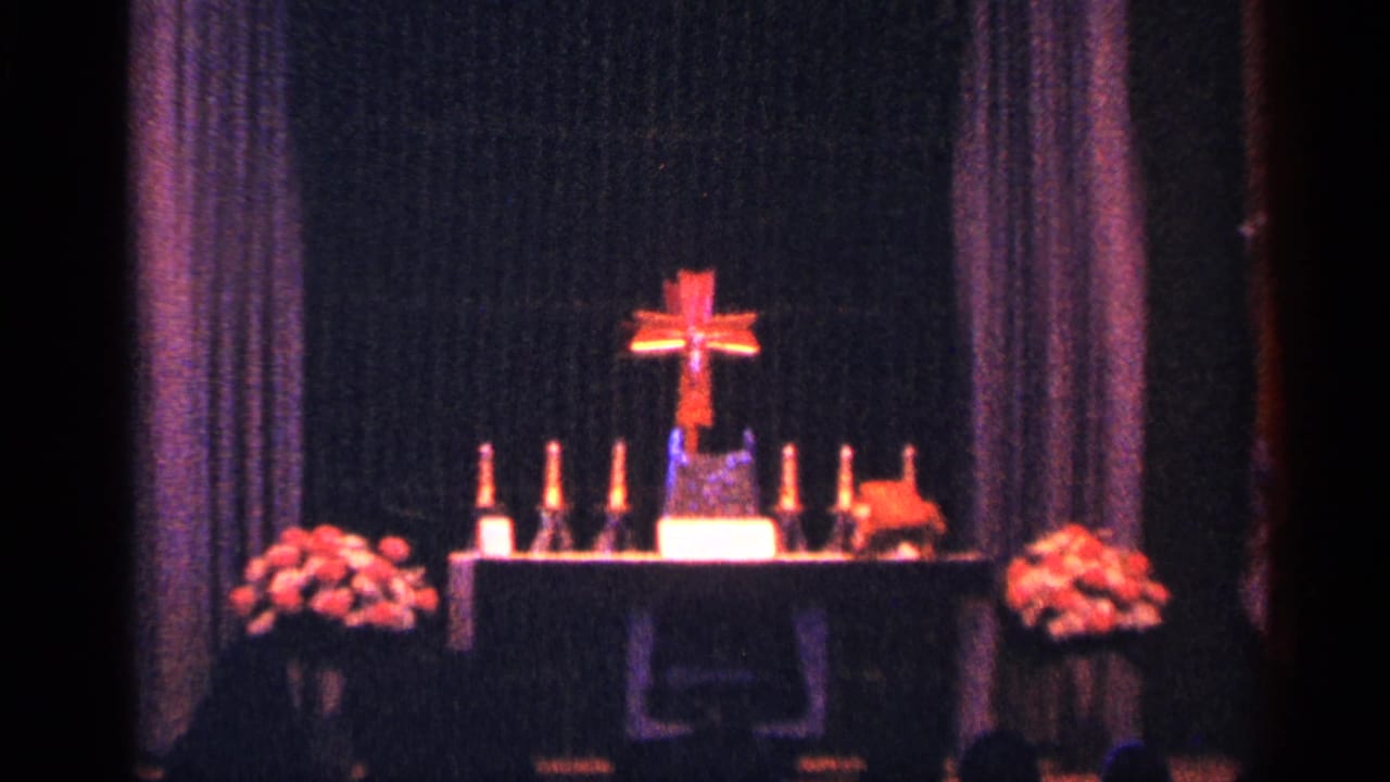 Church altar with red cross, candles, flowers, and dark drapery.