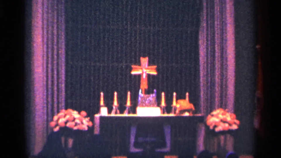 1964 New York City home-movie frame showing a wider view of the altar with cross, candles, flowers, and purple-toned drapery.