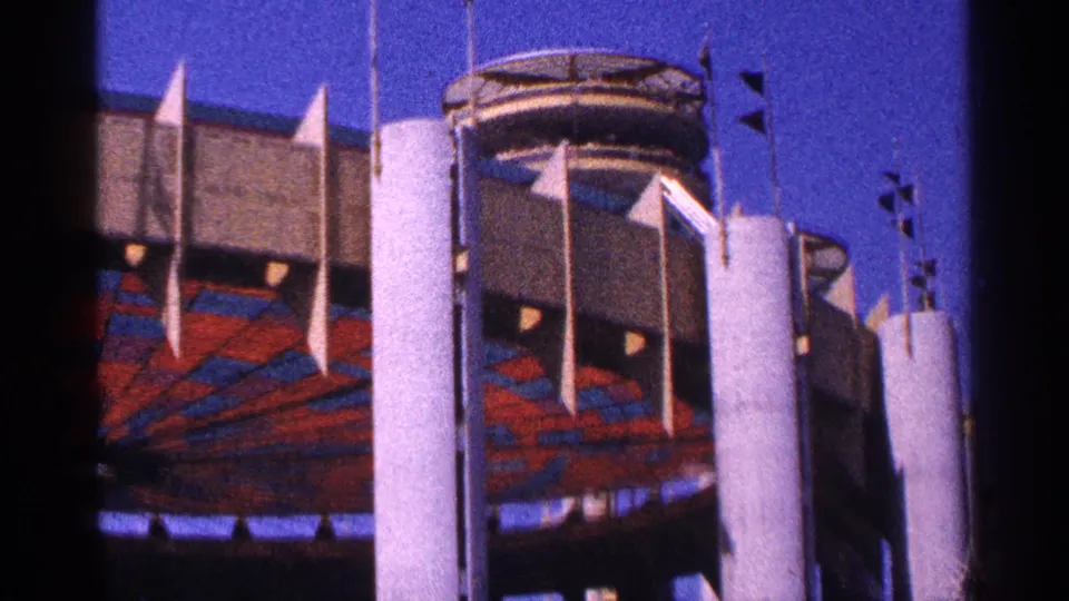 1964 New York City frame of a large modern structure with tall white columns, flags, and patterned red-blue exterior surfaces.