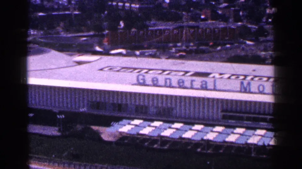 1964 New York World's Fair frame with the General Motors Pavilion sign, long facade, walkway area, and rows of parked cars below.
