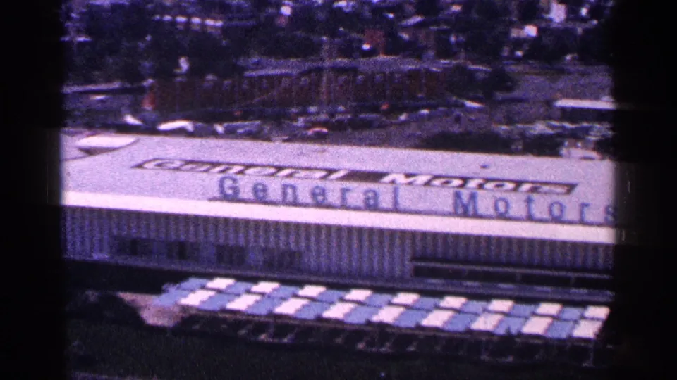 1964 New York World's Fair view of the General Motors Pavilion from above, showing the roof, large sign, paths, and parked cars nearby.
