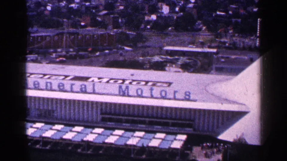 Elevated 1964 New York World's Fair frame showing the General Motors pavilion roof sign, canopy edge, trees, roads, and tiny visitors nearby.