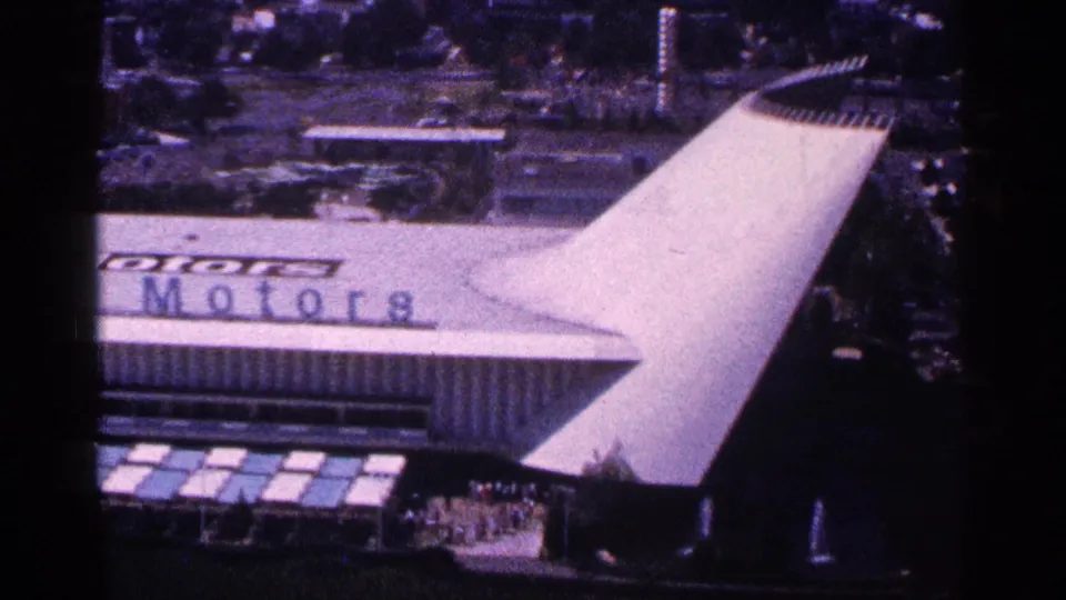Elevated frame from 1964 New York World's Fair footage with the General Motors pavilion partly cropped, its white canopy, roof sign, and paths visible.