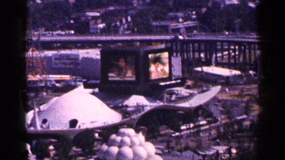 Elevated 1964 World's Fair view showing a white dome-like pavilion, curved paths, display screens, vehicles, and visitors below.