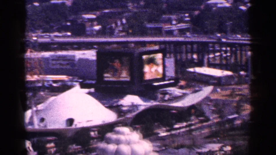 1964 New York World's Fair fairground from above, with domed pavilion forms, display screens, curved circulation paths, cars, and crowds.
