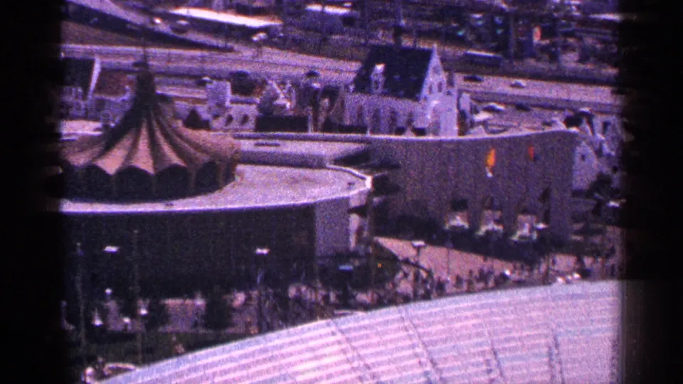 Elevated 1964 World's Fair view showing a dark tent-like roof, a large curved pavilion, roadway traffic, paths, and visitors.