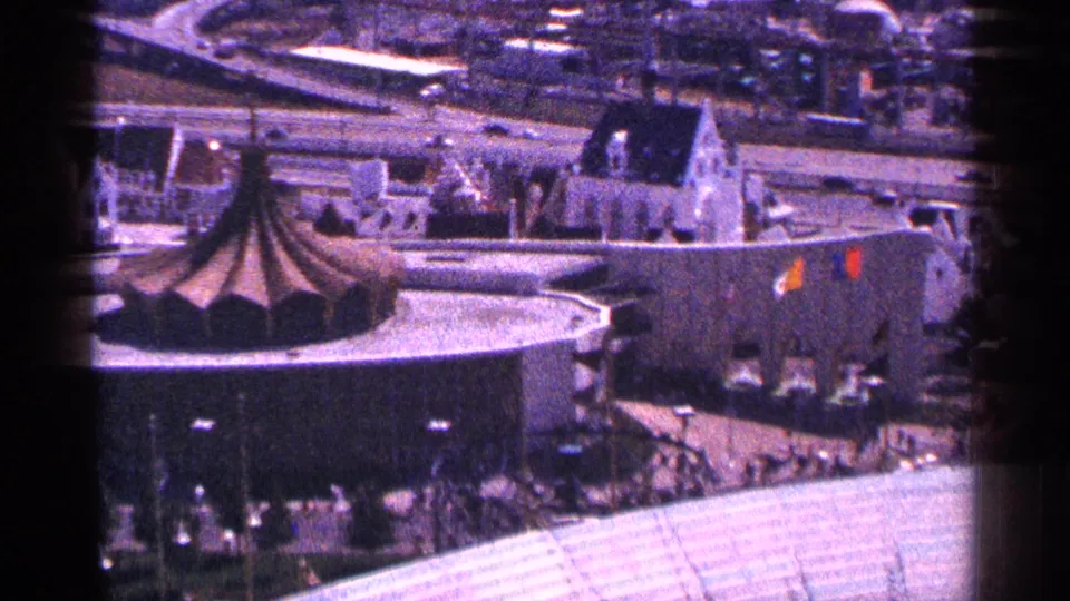 1964 New York City home-movie frame with a round conical-roof building, surrounding streets, rooftops, and soft purple film color.