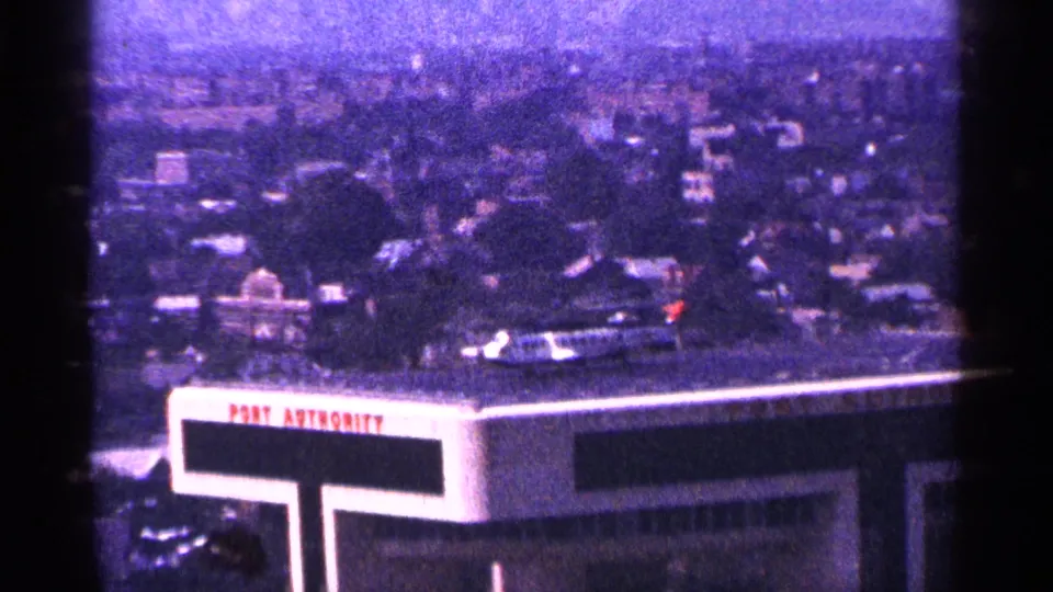 1964 New York City frame showing Port Authority rooftop lettering, nearby traffic, trees, and distant buildings under a hazy sky.