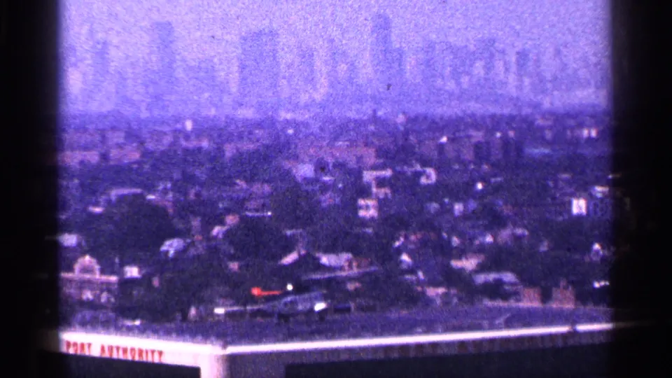 1964 New York City frame showing a wider rooftop helicopter view, Port Authority lettering, and a hazy skyline on purple-tinted film.