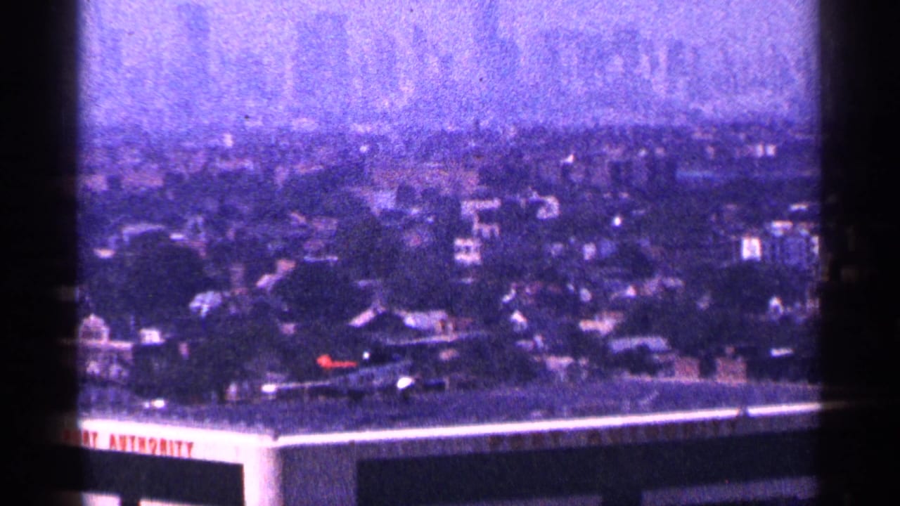 1964 New York City home-movie frame showing rooftops through haze with partial Port Authority signage at the lower edge.