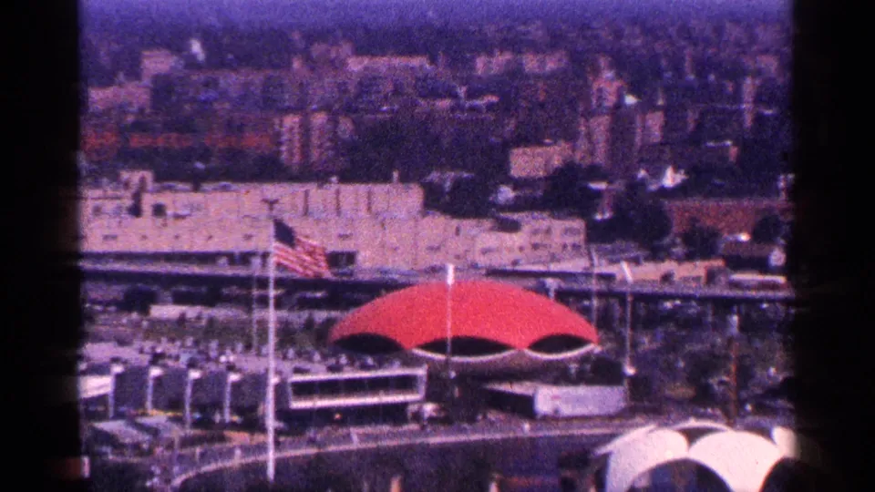 1964 New York City aerial home-movie view with rooftops, an American flag, and a distinctive red arched roof in the foreground.