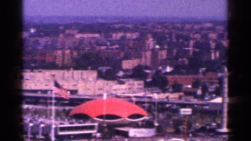 The elevated New York City view continues, with the red dome-like roof, American flag, and distant buildings softened by haze and 8mm grain.