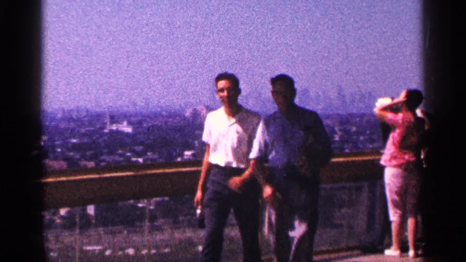 On a New York City observation deck in 1964, two adult men pass the railing as a woman in a pink top aims a camera toward the skyline.