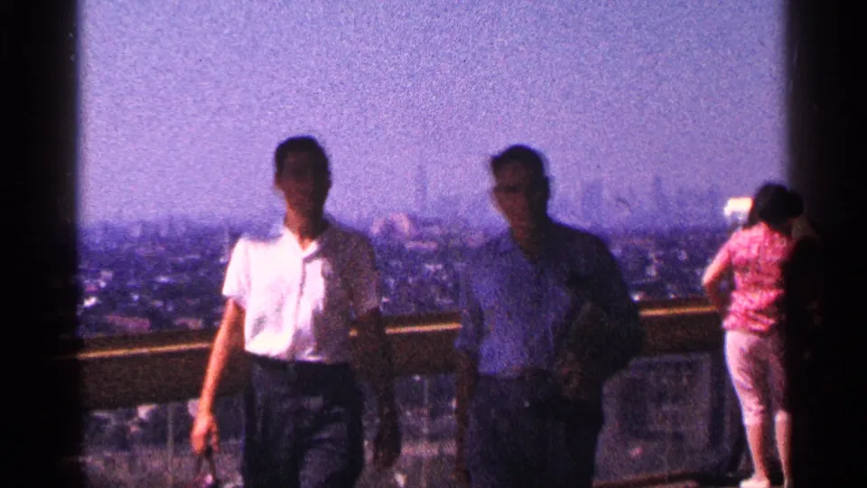 Adult visitors on the 1964 New York City observation deck are silhouetted by bright light, with the skyline visible beyond the railing.