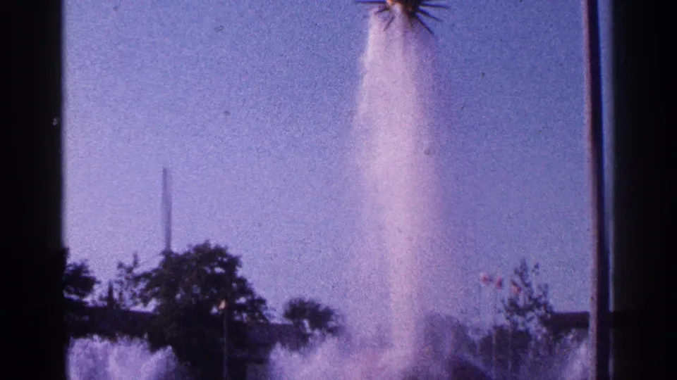 1964 New York City 8mm view of a high fountain plume, trees, and dark spectator silhouettes at the edge of the frame.