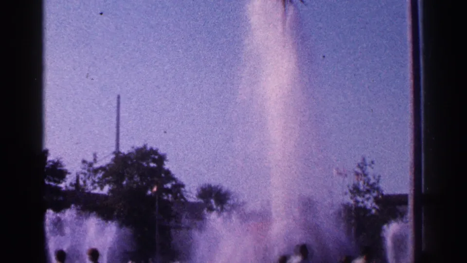 Visitor-shot 1964 New York City frame showing fountain spray, trees, and silhouetted people watching from the foreground.