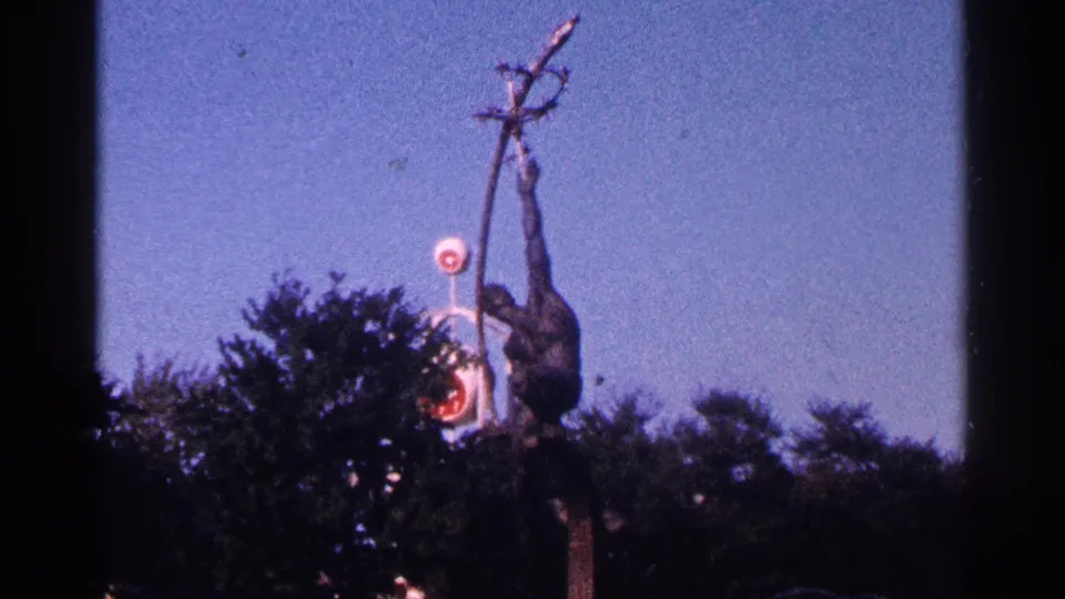 1964 New York City 8mm frame showing the primate-like sculpture more centrally, with trees below and a small round sign behind it.