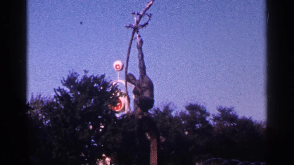 1964 New York City 8mm frame showing the dark primate-like statue, raised form, blue sky, tree canopy, and round red-and-white sign.