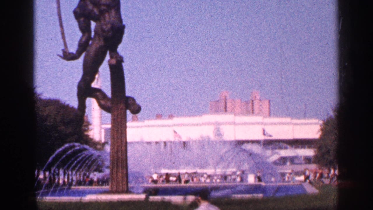 Bronze statue on a pedestal beside fountain jets and a white building.