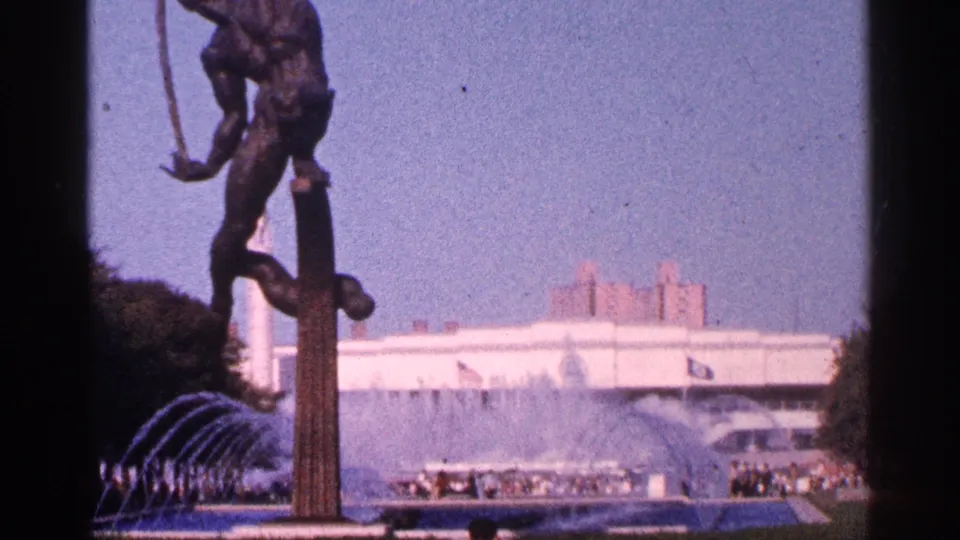 The frame holds on the plaza statue and active fountain, with small groups of visitors and a modern white building behind it in New York City.