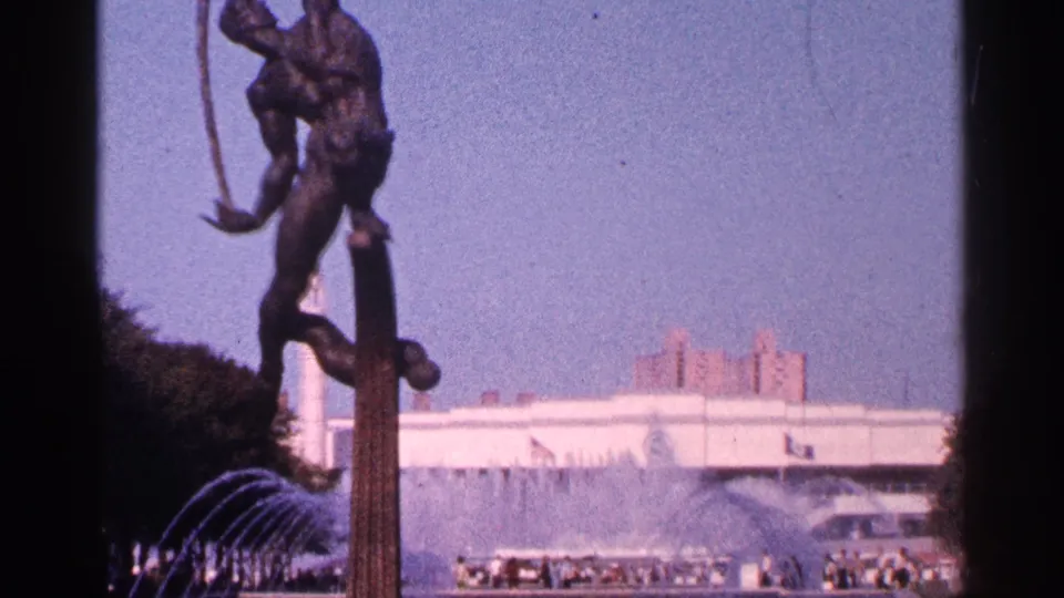 A soft 8mm view of the bronze statue and fountain arcs, with people seated or standing along the plaza and buildings in the distance.