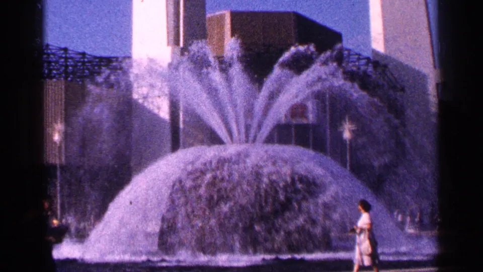 A large public fountain sprays in multiple arcs while a person stands near the edge, with modern buildings and plaza features behind it.