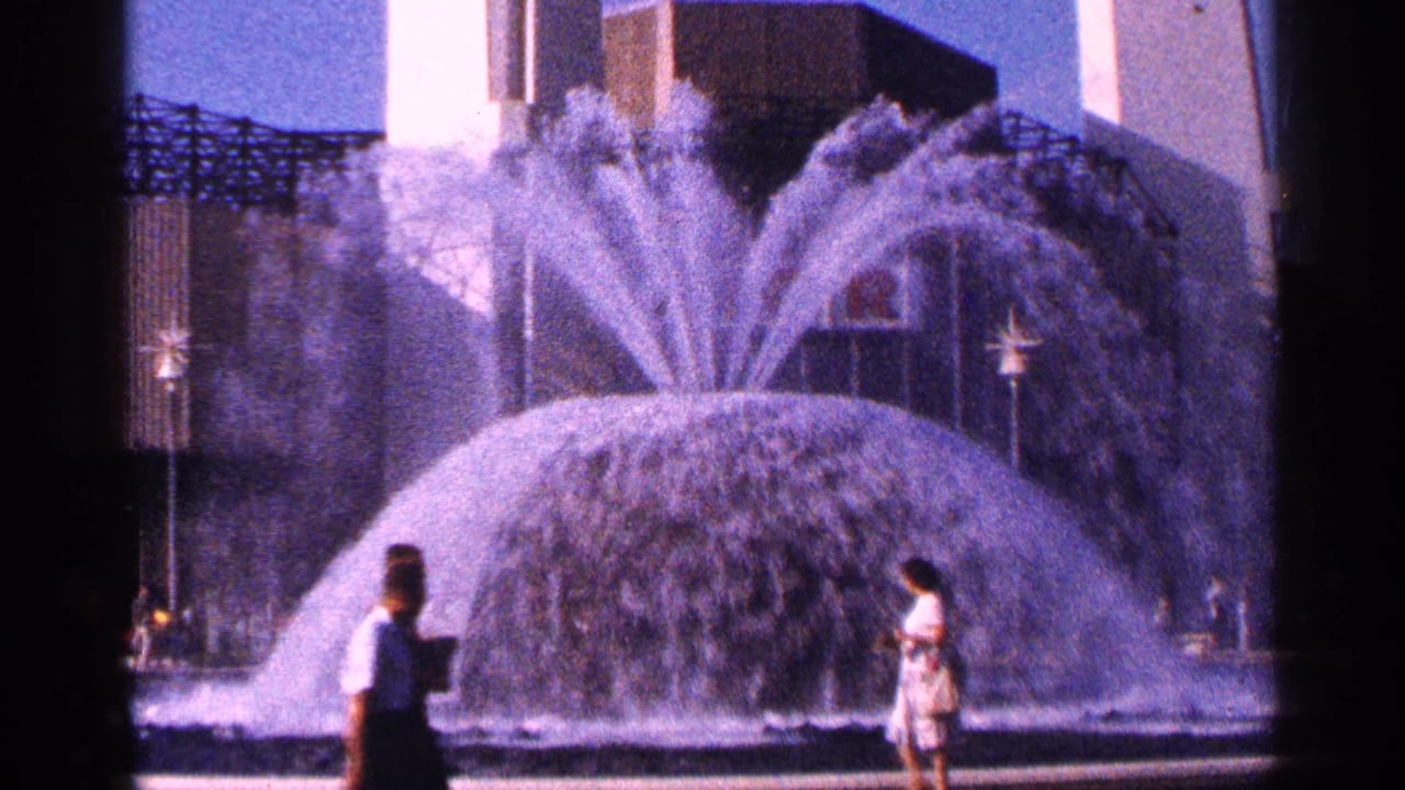 1964 New York City home-movie frame showing a large active fountain with pedestrians crossing the foreground.