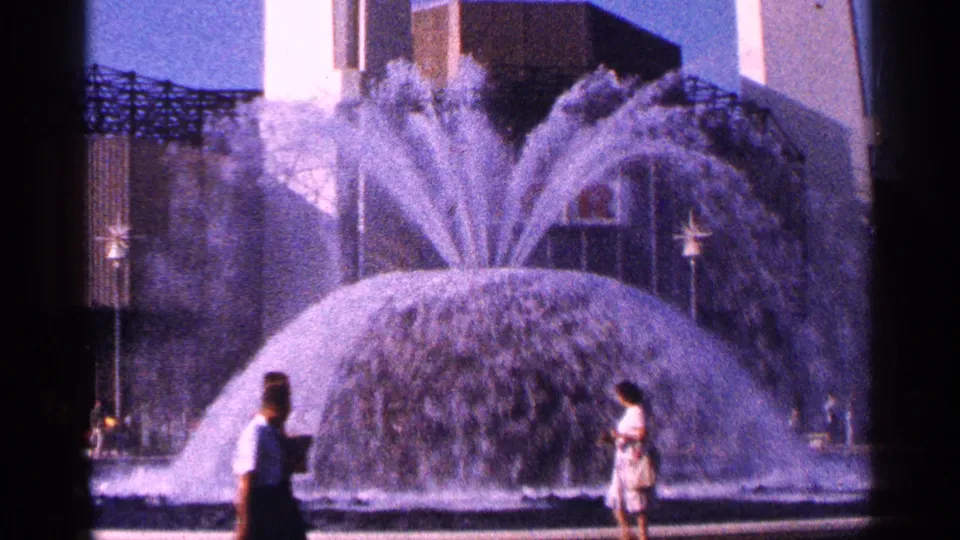 1964 New York City home-movie frame showing a large active fountain with pedestrians crossing the foreground.