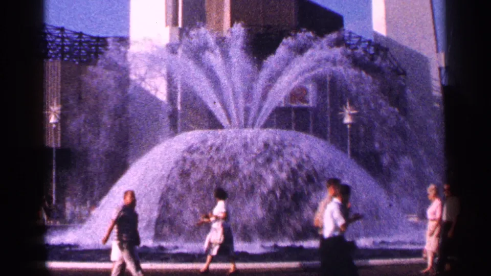 1964 New York City frame with several pedestrians moving past a high-spraying public fountain and modern building backdrop.