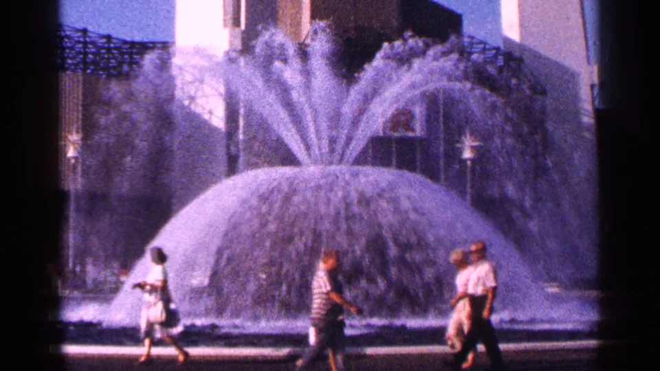 Home-movie view of a 1964 New York City fountain scene, with pedestrians passing beneath a modern building facade.