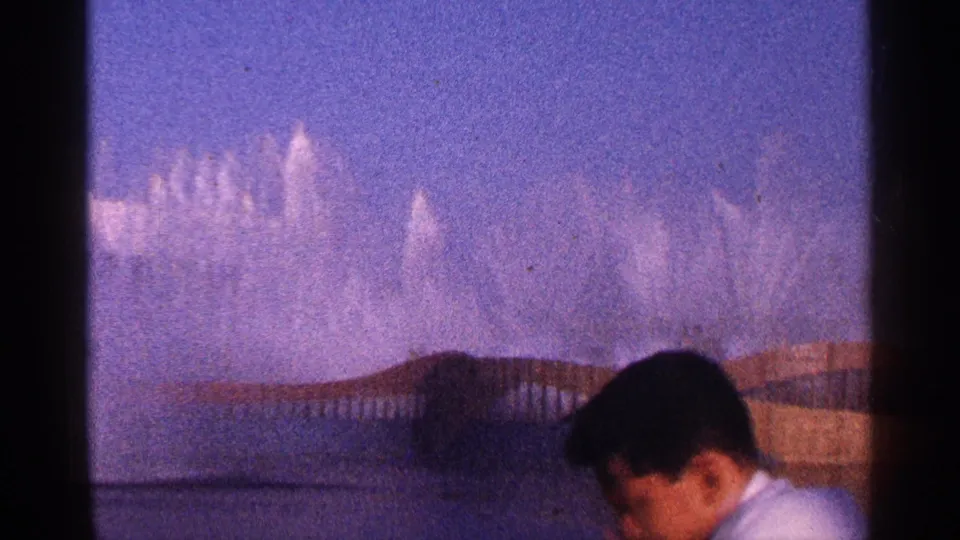 1964 New York City home-movie frame with a young man in a white shirt crossing below the distant fountain jets.