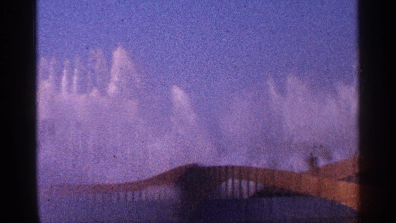 Fountain jets arc above a curved foreground structure under a blue sky.