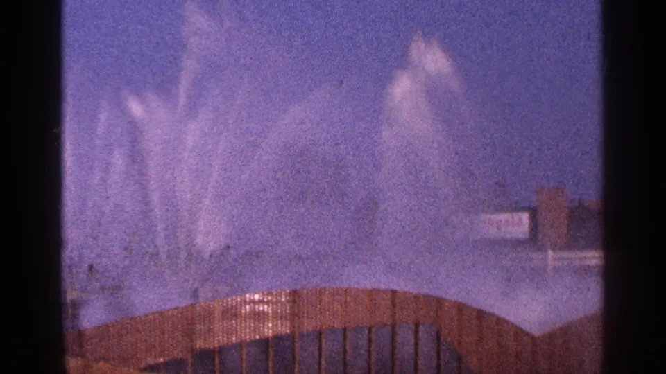 1964 New York City 8mm frame with a high fountain burst, curved foreground structure, and soft distant building forms.