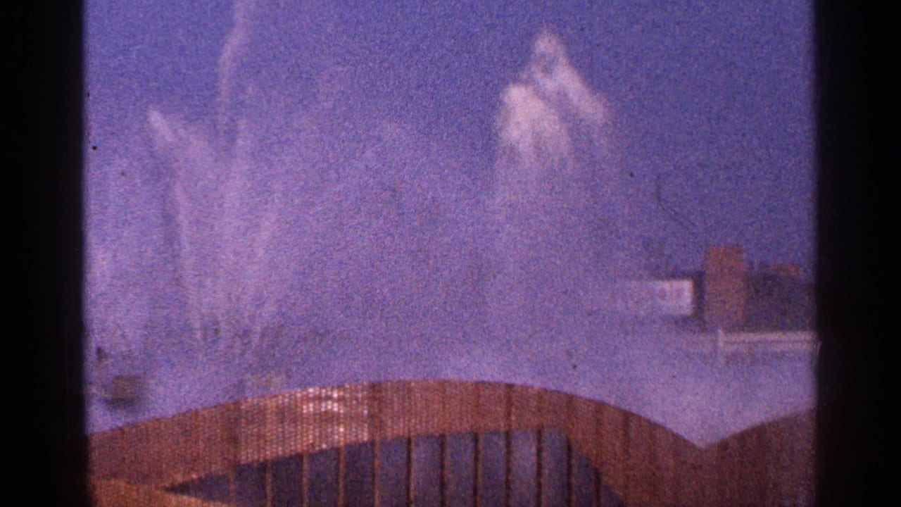 1964 New York City 8mm frame with tall fountain jets behind a curved brown railing and hazy blue sky.