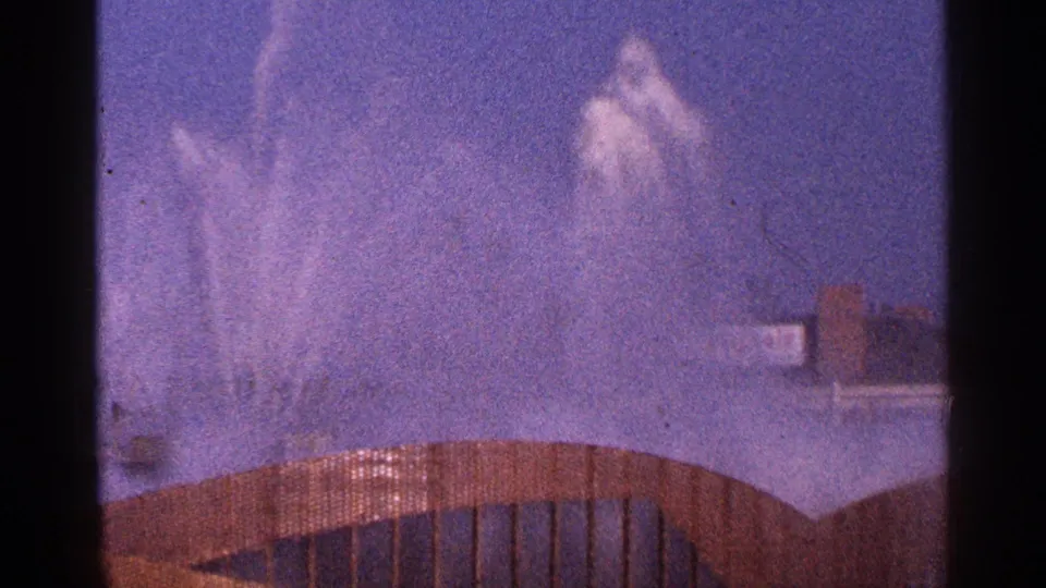 1964 New York City 8mm frame with tall fountain jets behind a curved brown railing and hazy blue sky.