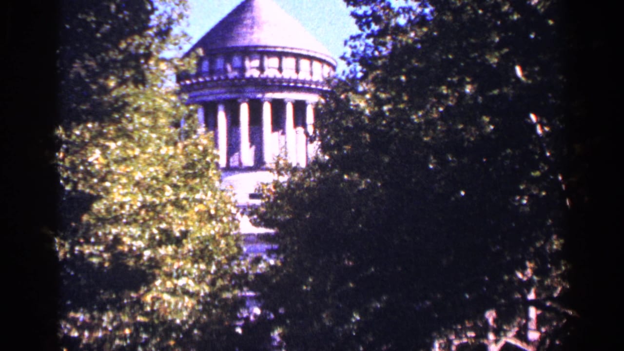 Domed stone building with columns partly hidden behind green trees.