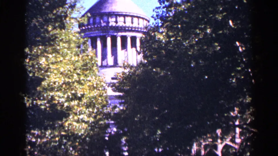 1964 New York City 8mm frame with a domed stone building viewed through bright green leaves and deep shade.