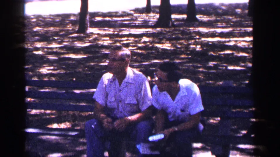 Two men sit on a park bench in 1964 New York City footage, partly shaded by trees and facing toward activity off camera.