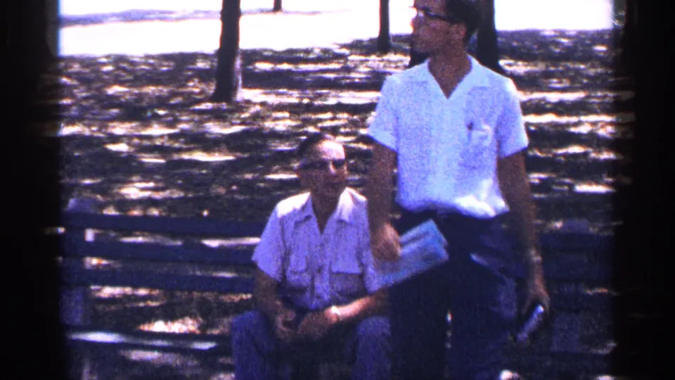 In a 1964 New York City home-movie frame, one man remains seated on a bench while another stands nearby in a light shirt.