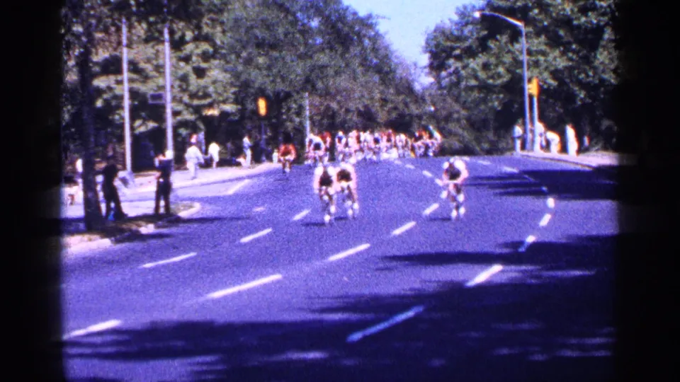 Bicycle racers move down a broad New York City street in 1964, with trees, traffic signals, and spectators visible along the route.