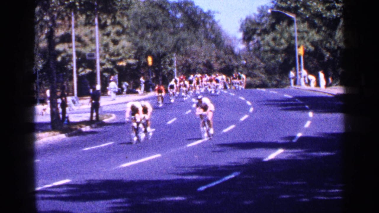 Cyclists ride around a broad city-street curve with trees, traffic signals, and sidewalk spectators nearby.