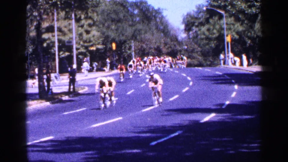 In 1964 New York City footage, cyclists approach and round a sunlit street curve while spectators stand along the sidewalk.